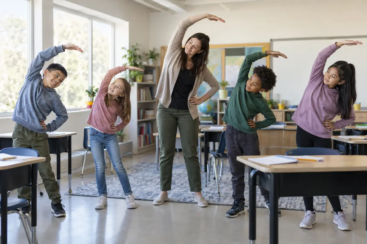 A teacher and four elementary students doing a gentle standing side stretch beside classroom desks in natural window light.